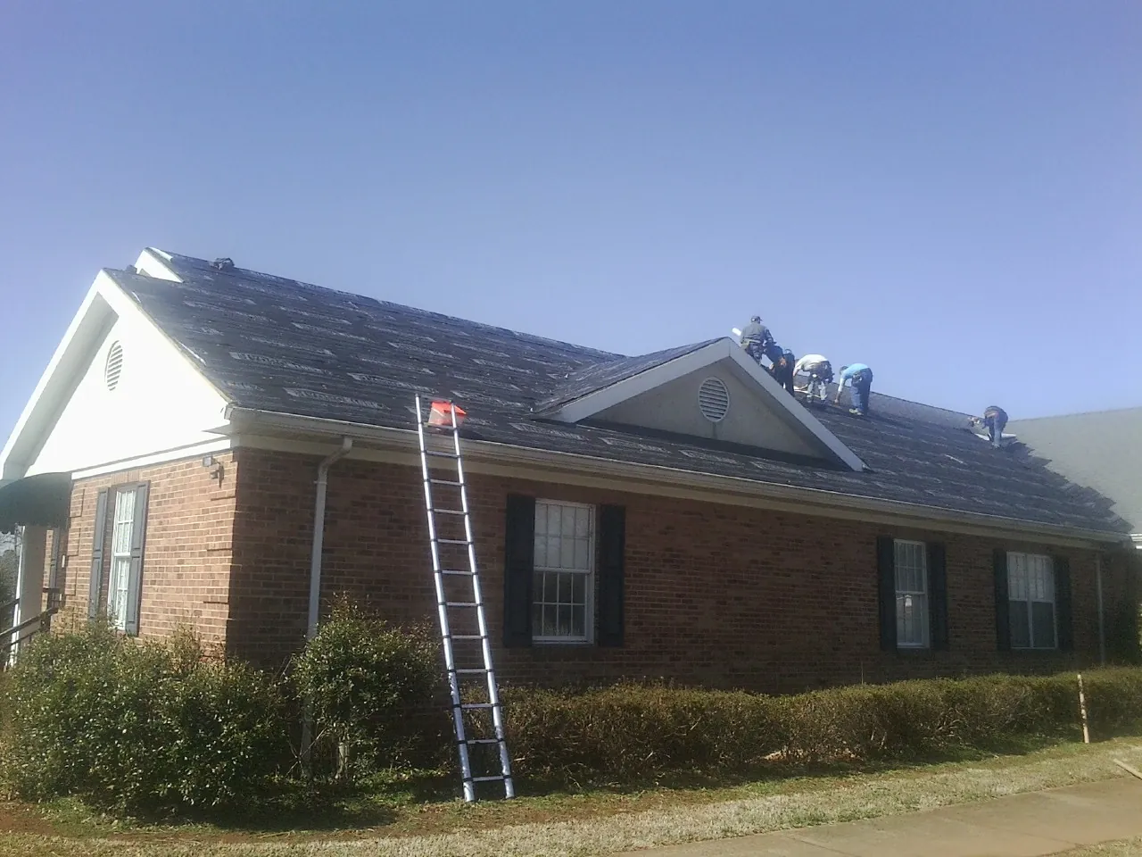 Crew tearing shingles off a brick ranch roof with a ladder set — Emergency Roof Repair work by our crew in Pineville